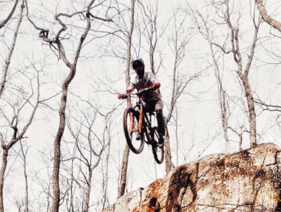 A man going over a jump on a mountain bike in the woods (Instagram@weycyde)